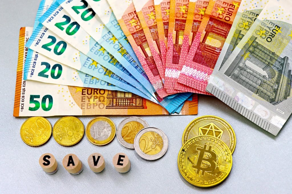 Euro banknotes and cryptocurrency coins arranged on a white surface with blocks spelling 'SAVE'.