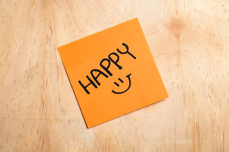 Orange sticky note on wood with the word 'HAPPY' and a smiley face.