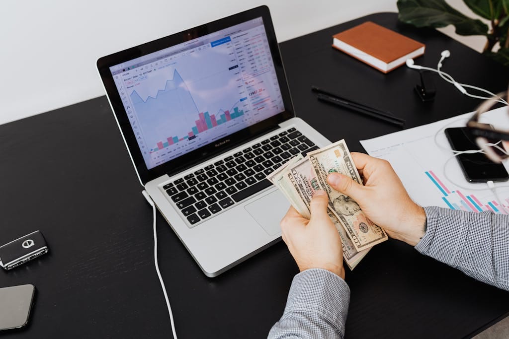 Person counting dollar bills with financial graphs on a laptop screen, symbolizing financial analysis and planning.
