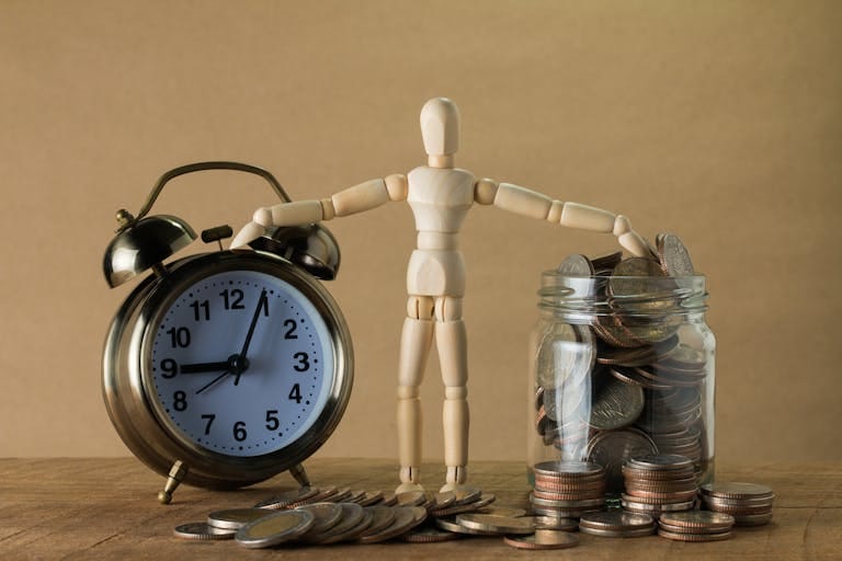 Wooden mannequin holding an alarm clock and a jar full of coins, symbolizing time and money.