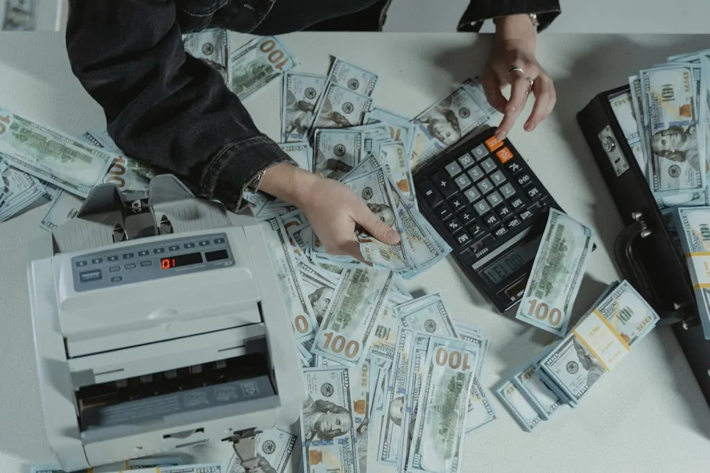 18 Simple Money Hacks That Add Up Fast 1 Hands counting 100 dollar bills using a calculator and money counter on a table.