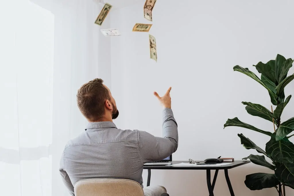 5 Money Hacks You Need in 2026 1 Man throwing cash in the air while sitting at a desk indoors, symbolizing financial success.
