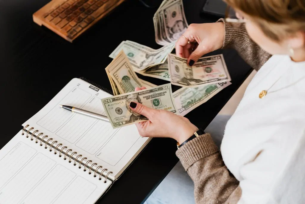 Budgeting Hacks to Save Money Fast 1 Woman organizing finances, counting dollar bills at desk with open notebook.