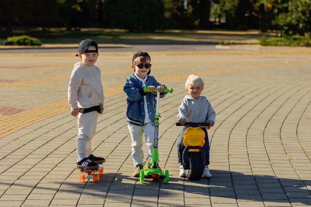 Three young children play outside with a skateboard, scooter, and toy bike on a sunny day.